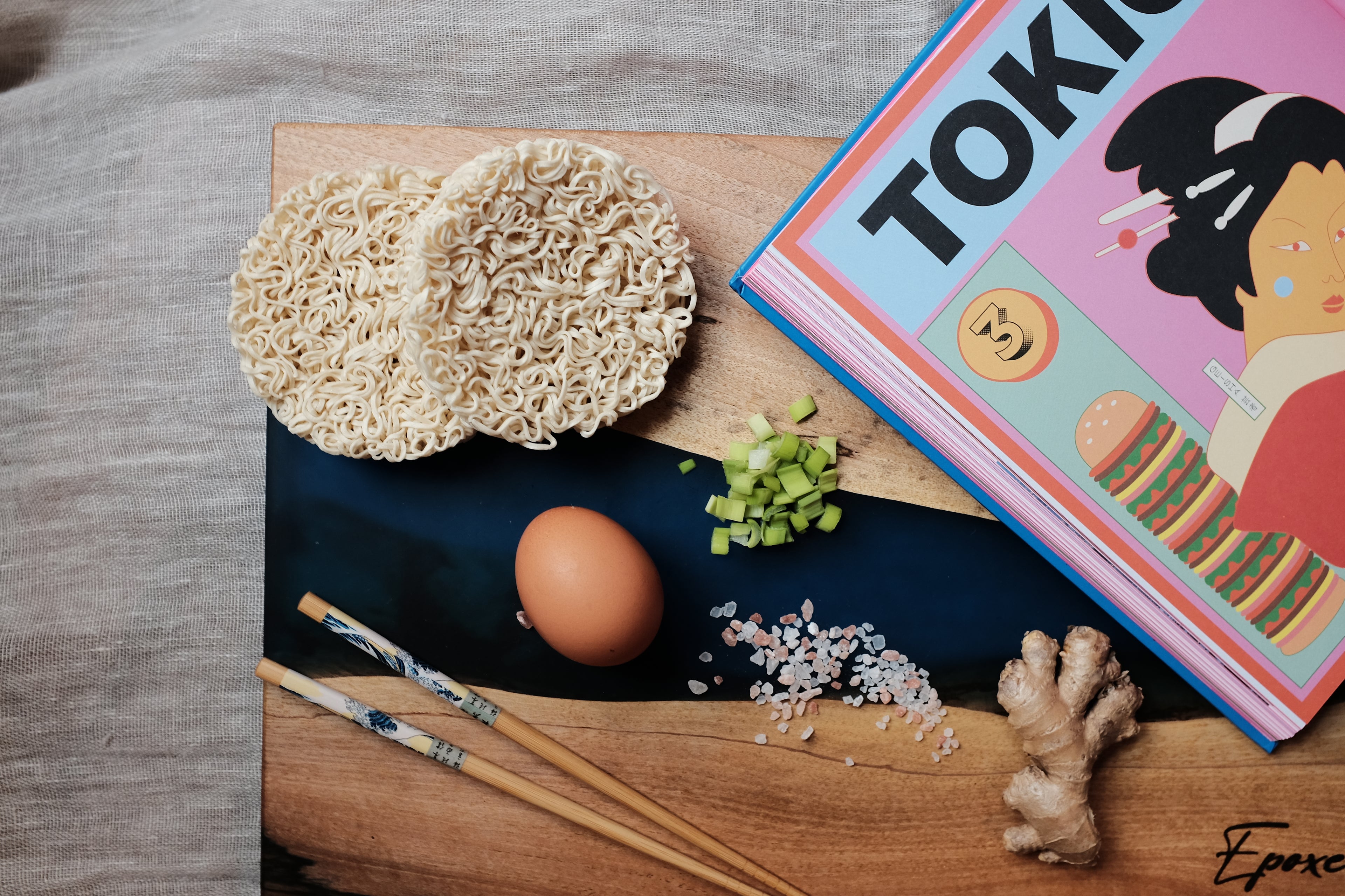Epoxy blue wooden board adorned with a delightful arrangement of noodles, eggs, chopsticks, and a Tokyo street food coffee table book - an enticing fusion of aesthetics and gastronomy, capturing the essence of vibrant culinary culture in Tokyo
